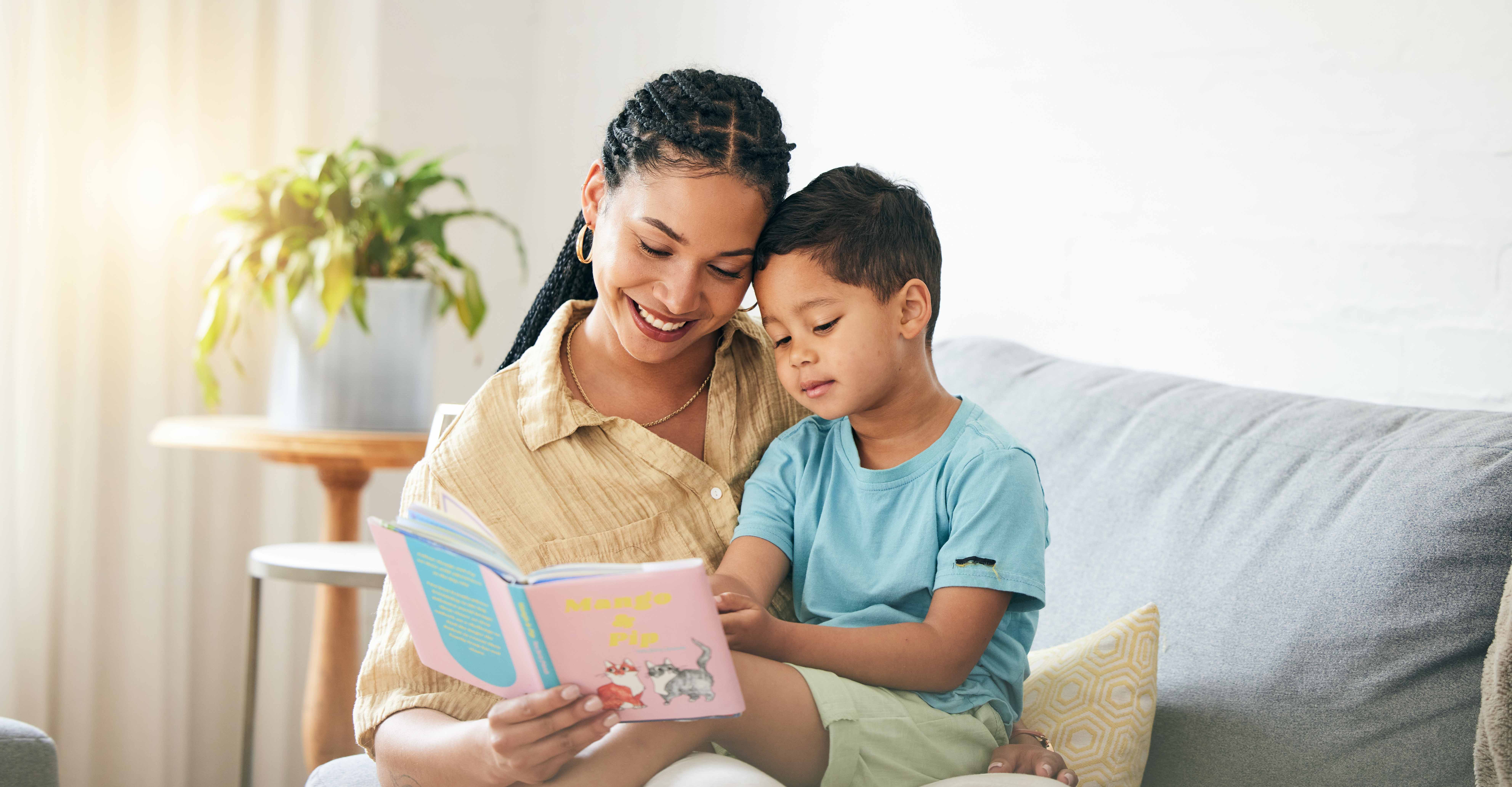 A mother sitting on a sofa with her young child on her lap, reading a book to him.